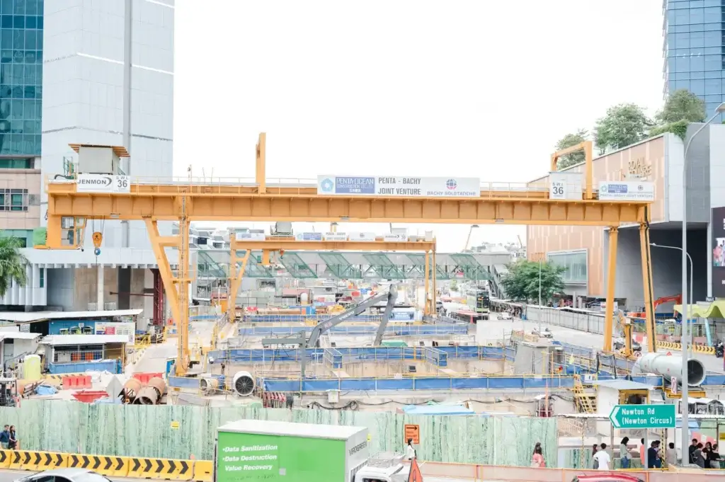 Large-scale MRT construction site in Singapore featuring heavy-duty gantry cranes, excavation works, construction machinery, safety barriers, and underground infrastructure development along a busy urban roadway