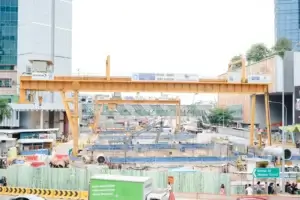 Large-scale MRT construction site in Singapore featuring heavy-duty gantry cranes, excavation works, construction machinery, safety barriers, and underground infrastructure development along a busy urban roadway