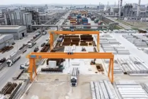 Two large bright orange gantry cranes stand over an active concrete yard filled with stacked construction materials, steel reinforcement bars, and precast concrete beams.